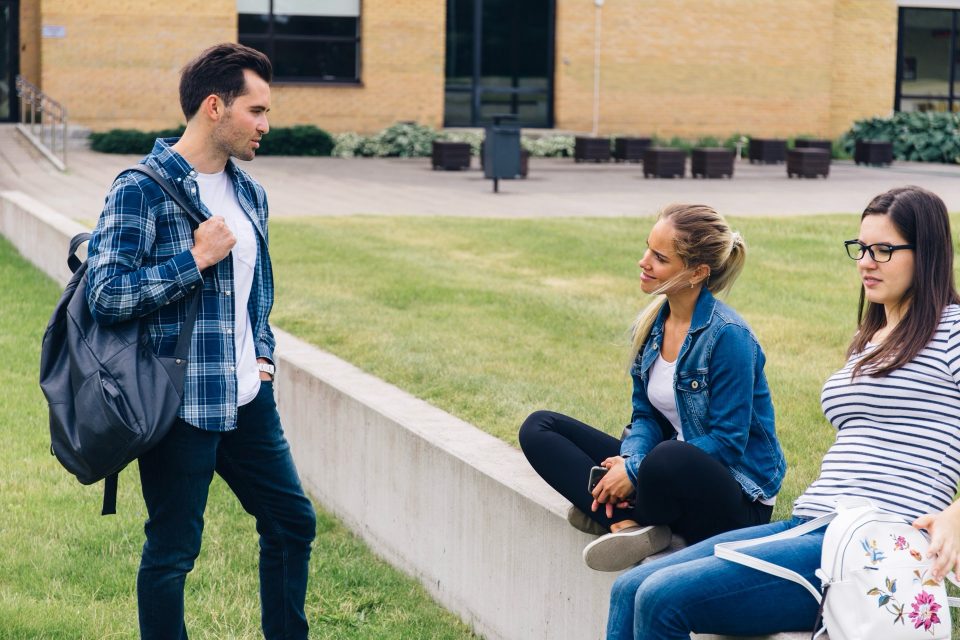 Tre studenter samtalar på en gräsmatta utanför ett tegelhus på campus