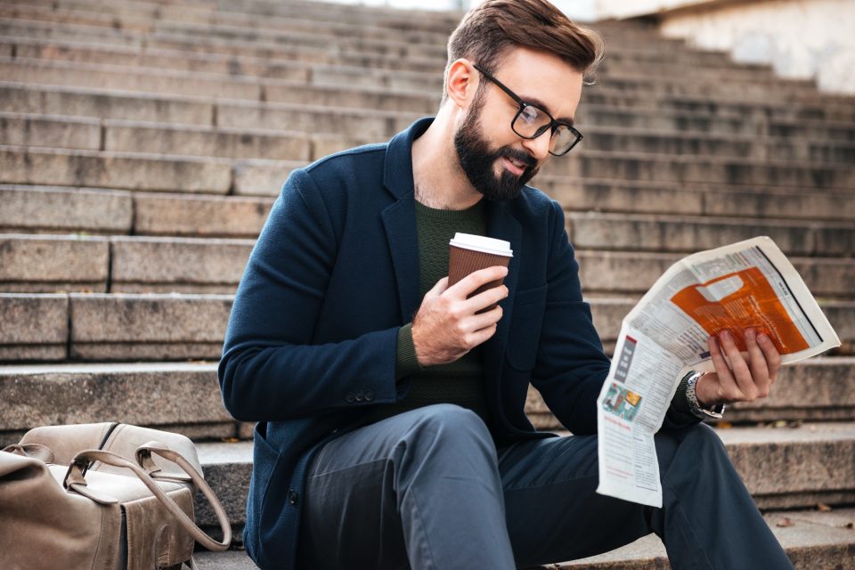 Ung man med kaffe läser en tidning på en trappa utomhus